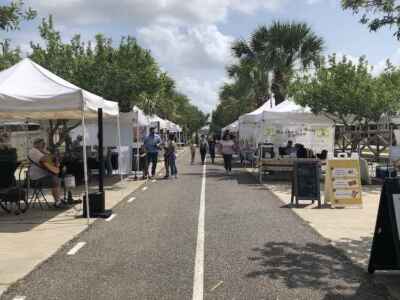 Two rows of white tents are lined up with shippers walking between the rows. The market is in a park, which is lined by trees and grassy fields.